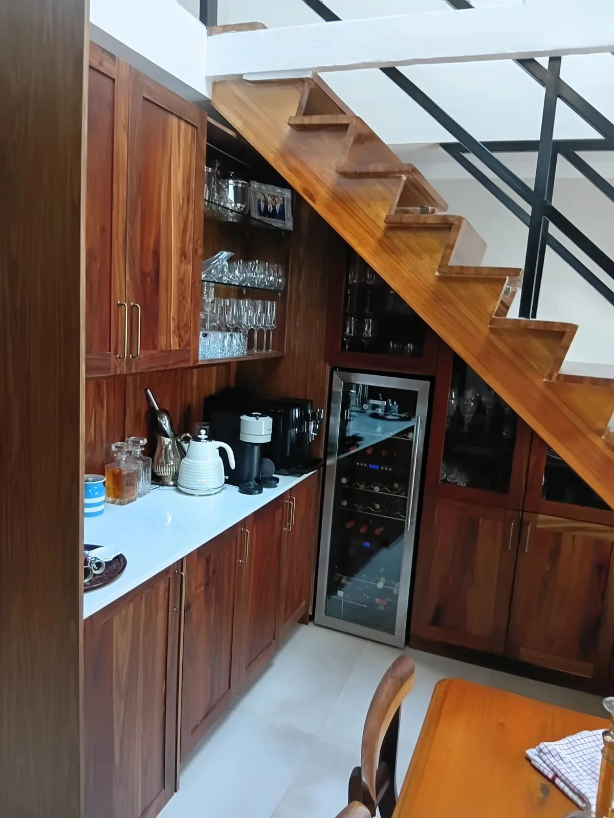 Under-Stair Bar, Full Run — The full run of the under-stair bar: tall cupboards on the left, the coffee station and wine fridge in the middle, and glazed display cabinets stepping down with the underside of the stair.