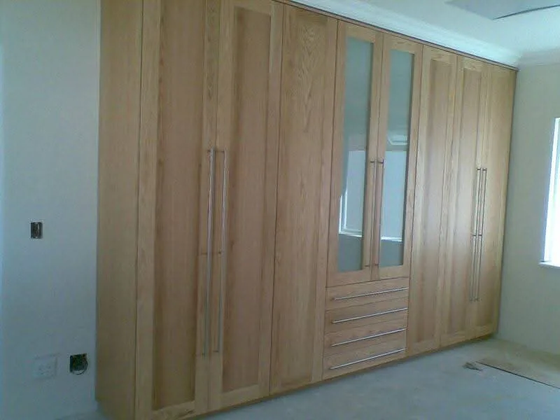 Solid Oak Built-In Wardrobe — Floor-to-ceiling oak wardrobes with a shaker profile, a mirrored centre section and a four-drawer bank. Long brushed-steel handles keep the vertical lines running clean.
